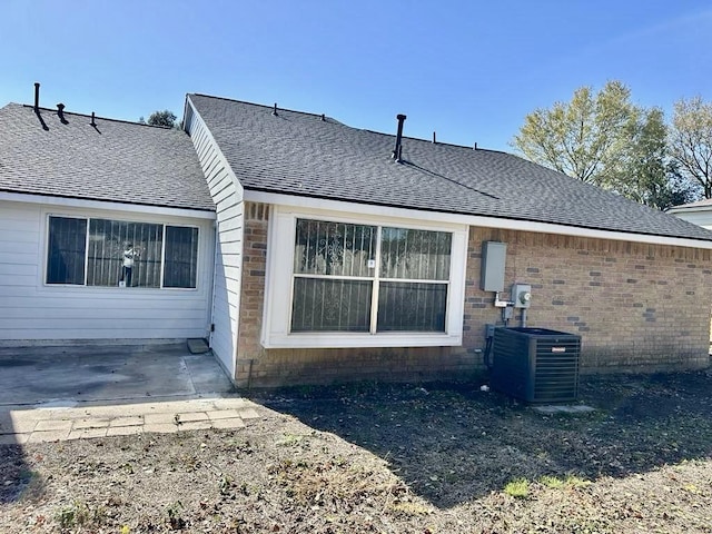 rear view of property with a shingled roof, brick siding, central AC, and a patio
