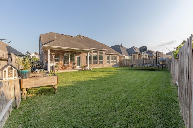 rear view of house with a trampoline, a fenced backyard, a yard, brick siding, and roof with shingles