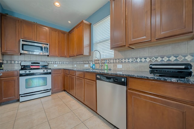 kitchen with brown cabinets, a sink, dark stone countertops, stainless steel appliances, and light tile patterned floors