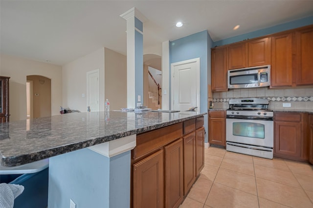 kitchen featuring appliances with stainless steel finishes, arched walkways, brown cabinets, light tile patterned floors, and dark stone countertops