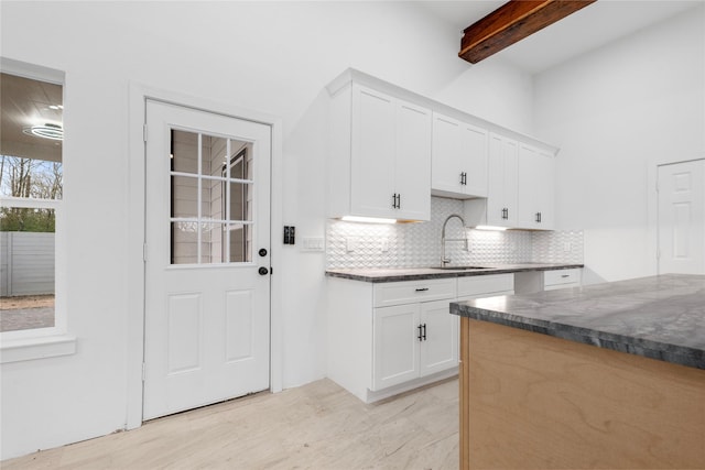 kitchen with a sink, beam ceiling, white cabinetry, dark stone counters, and backsplash