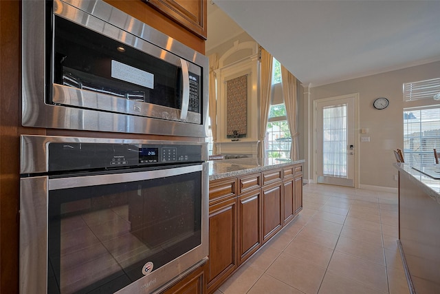 kitchen featuring light tile patterned floors, baseboards, appliances with stainless steel finishes, brown cabinets, and light stone countertops