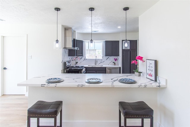 kitchen with wall chimney range hood, a sink, stainless steel appliances, a peninsula, and a kitchen bar