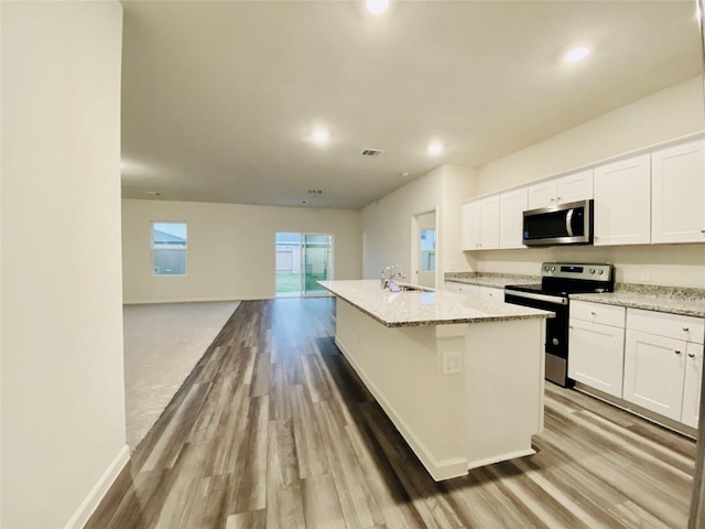 kitchen featuring an island with sink, white cabinetry, wood finished floors, stainless steel appliances, and light stone counters