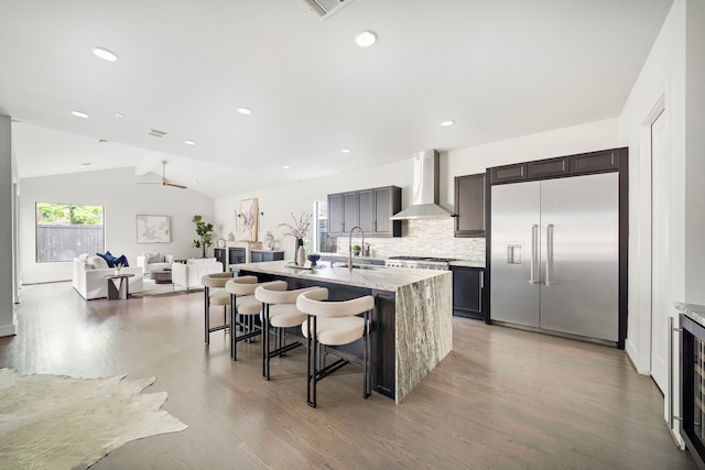 kitchen with a breakfast bar area, a center island with sink, wall chimney exhaust hood, light wood finished floors, and stainless steel built in refrigerator