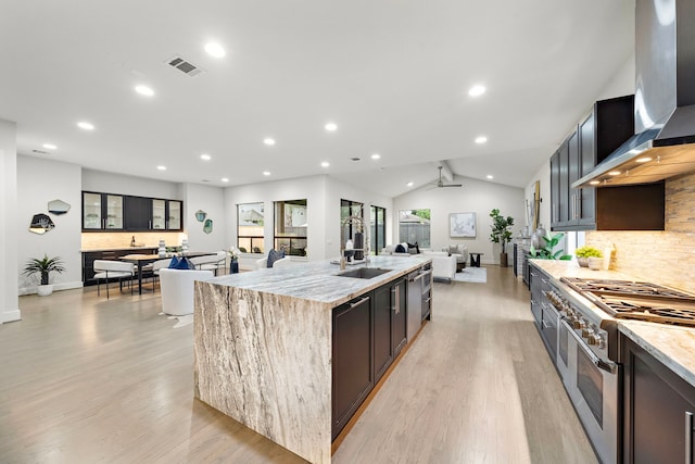 kitchen featuring wall chimney range hood, double oven range, visible vents, light stone countertops, and an island with sink