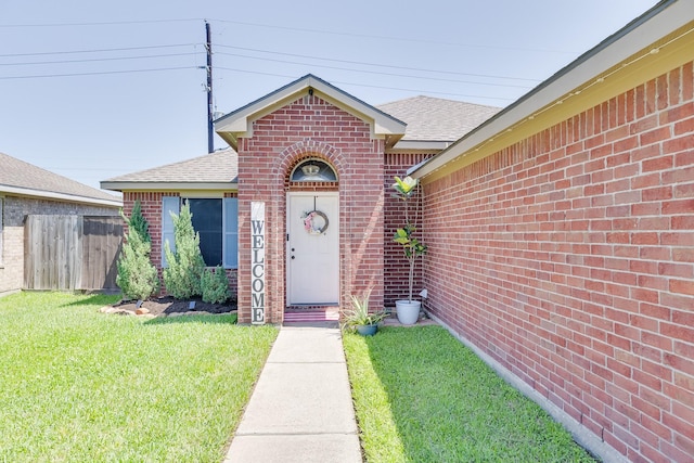 entrance to property featuring brick siding, fence, a lawn, and roof with shingles