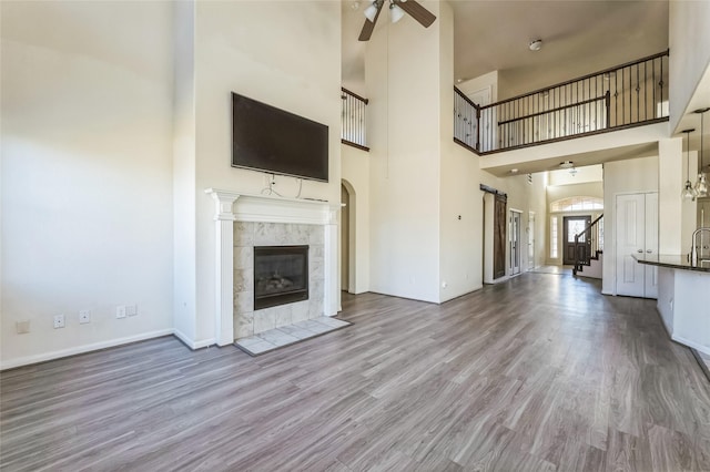 unfurnished living room featuring baseboards, a tiled fireplace, a ceiling fan, wood finished floors, and a towering ceiling