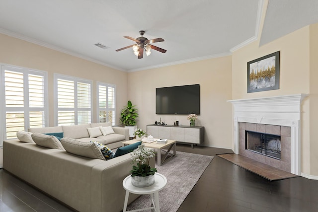 living room with visible vents, a tiled fireplace, ceiling fan, crown molding, and wood finished floors