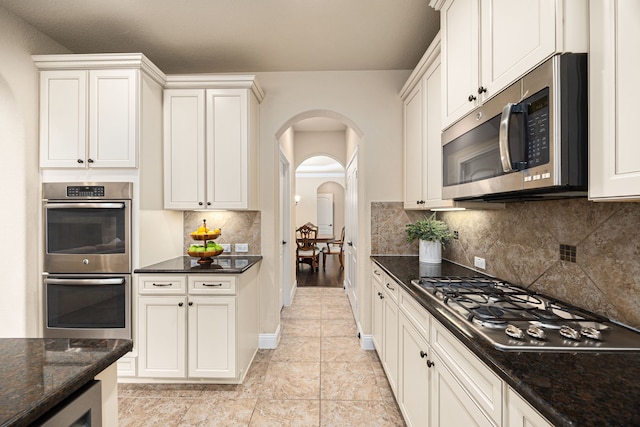 kitchen featuring decorative backsplash, arched walkways, light tile patterned floors, dark stone countertops, and stainless steel appliances