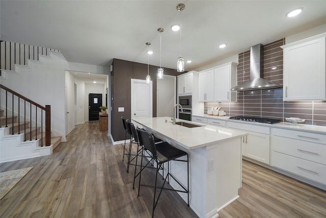 kitchen with a sink, wall chimney range hood, light wood-type flooring, a kitchen island with sink, and stainless steel appliances
