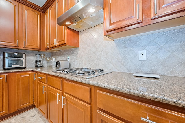 kitchen with light stone counters, light tile patterned flooring, under cabinet range hood, stainless steel gas cooktop, and brown cabinetry