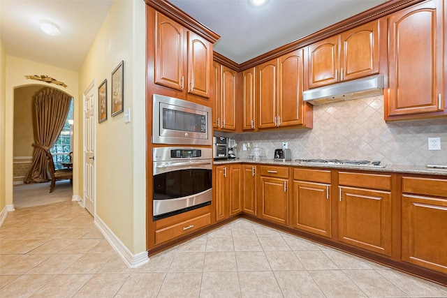 kitchen featuring light tile patterned floors, tasteful backsplash, appliances with stainless steel finishes, light stone counters, and under cabinet range hood
