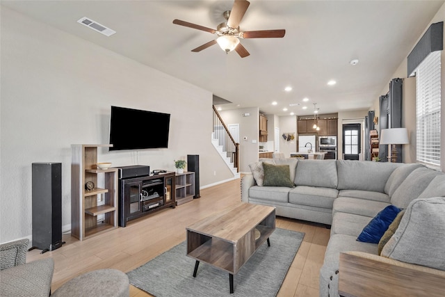 living room featuring a ceiling fan, visible vents, stairs, light wood finished floors, and baseboards