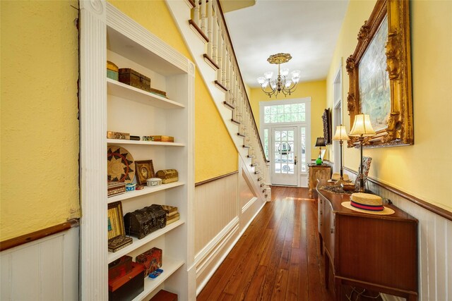 corridor with a wainscoted wall, stairs, an inviting chandelier, and dark wood-style floors