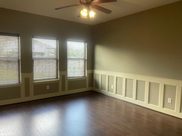 unfurnished room featuring a wainscoted wall, ceiling fan, dark wood-style floors, and a wealth of natural light