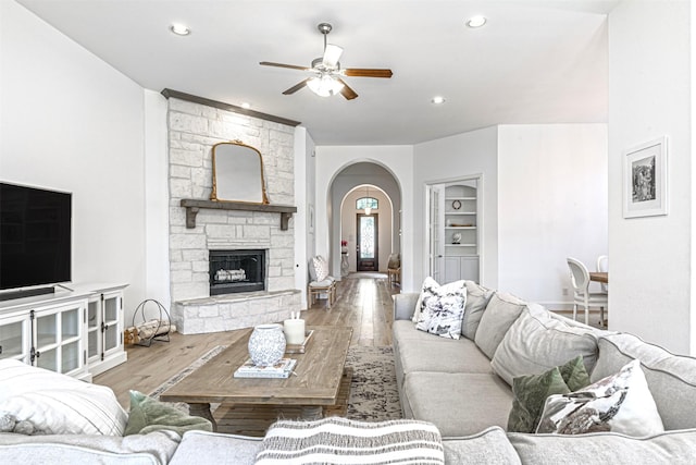 living room featuring a ceiling fan, a stone fireplace, wood finished floors, arched walkways, and baseboards