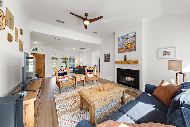 living area featuring light wood-type flooring, ceiling fan, crown molding, and a fireplace with raised hearth
