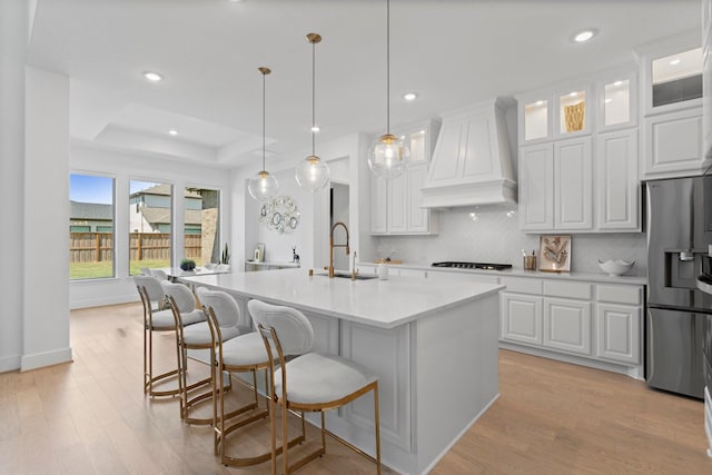 kitchen with light wood-type flooring, stainless steel refrigerator with ice dispenser, a sink, premium range hood, and decorative backsplash