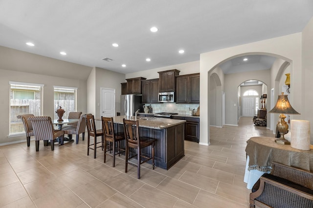 kitchen featuring dark brown cabinetry, a breakfast bar, a kitchen island with sink, decorative backsplash, and appliances with stainless steel finishes