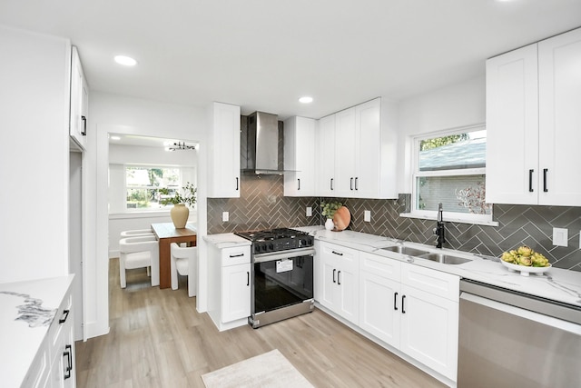 kitchen featuring stainless steel dishwasher, light wood-type flooring, wall chimney range hood, a sink, and gas range oven