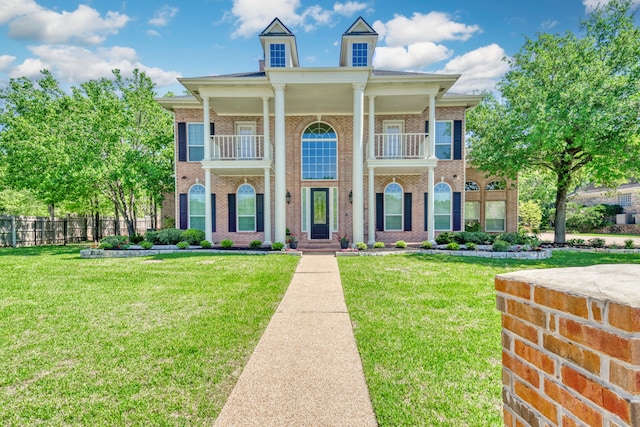 greek revival inspired property featuring a front yard, brick siding, a balcony, and fence