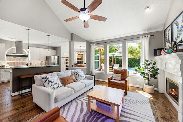 living room featuring high vaulted ceiling, a ceiling fan, a glass covered fireplace, baseboards, and dark wood-type flooring