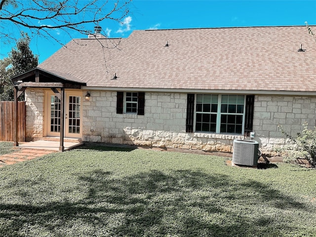 rear view of house featuring central AC unit, a shingled roof, a yard, stone siding, and fence