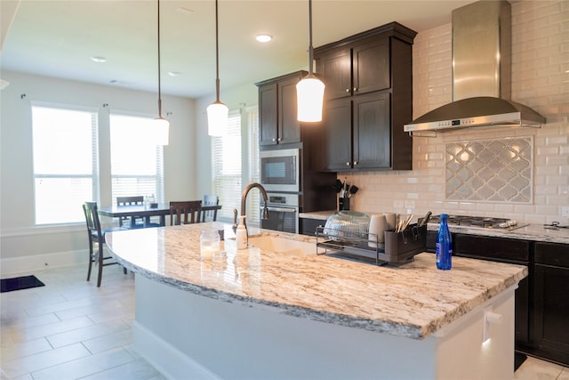 kitchen with tasteful backsplash, light stone countertops, a kitchen island with sink, baseboards, and wall chimney exhaust hood