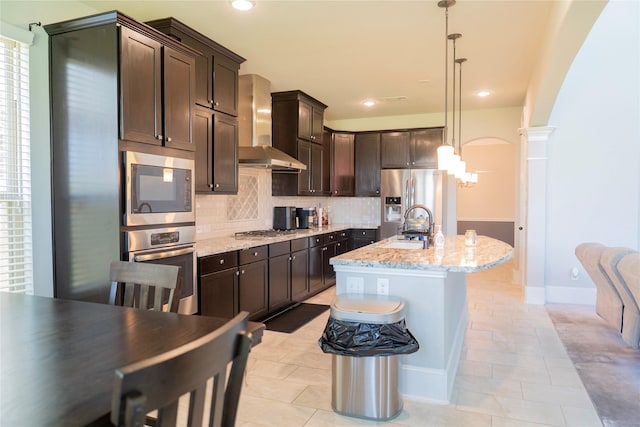 kitchen featuring wall chimney range hood, stainless steel appliances, dark brown cabinets, light stone countertops, and backsplash