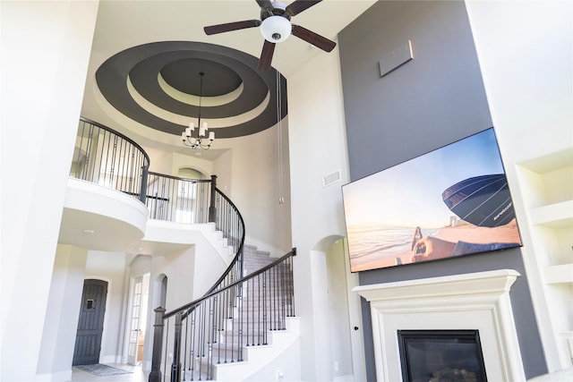 stairs with a glass covered fireplace, visible vents, a towering ceiling, arched walkways, and ceiling fan with notable chandelier