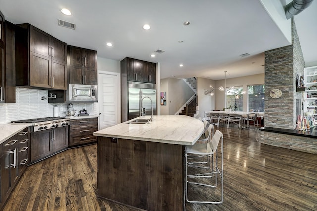 kitchen featuring visible vents, built in appliances, dark brown cabinetry, dark wood-style floors, and a center island with sink