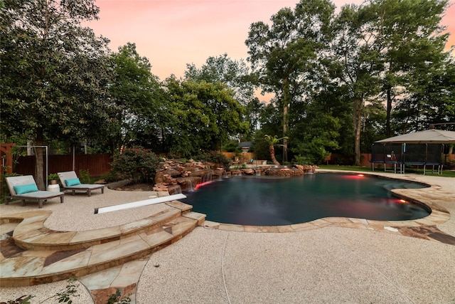 pool at dusk with a patio, fence, an outdoor pool, and a trampoline