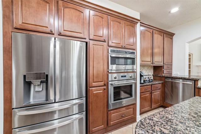 kitchen with brown cabinetry, dark stone countertops, stainless steel appliances, light tile patterned floors, and backsplash