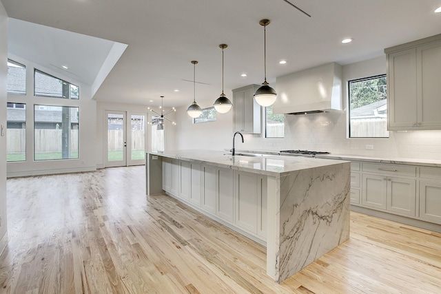 kitchen with custom range hood, light wood-style flooring, backsplash, light stone countertops, and a sink