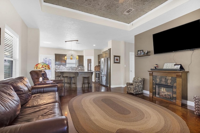 living room featuring a stone fireplace, visible vents, dark wood-style flooring, a raised ceiling, and baseboards