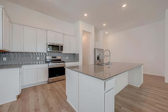 kitchen with a sink, appliances with stainless steel finishes, a kitchen island with sink, backsplash, and light wood-style flooring