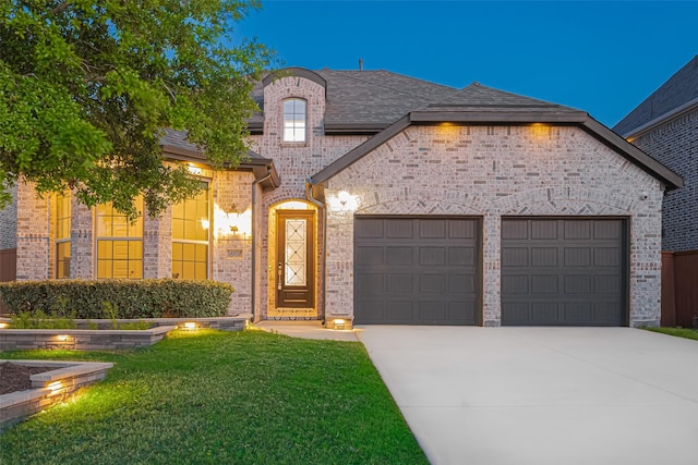 french provincial home with a front yard, a shingled roof, an attached garage, concrete driveway, and brick siding