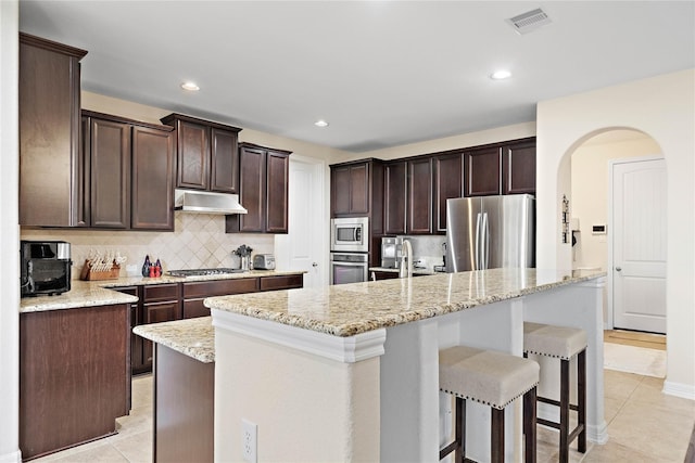 kitchen with light tile patterned floors, dark brown cabinetry, tasteful backsplash, under cabinet range hood, and stainless steel appliances