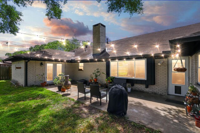 rear view of property with a shingled roof, brick siding, a chimney, a patio, and a yard