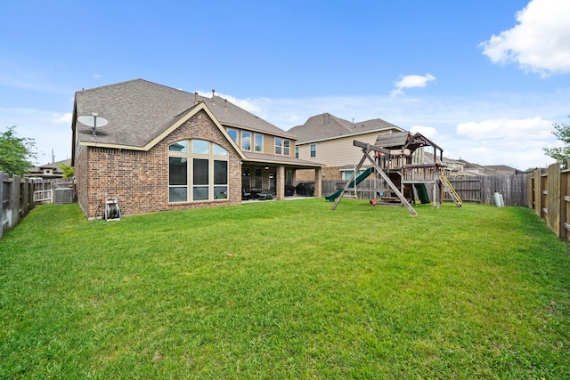 back of property with a playground, roof with shingles, a yard, brick siding, and a fenced backyard