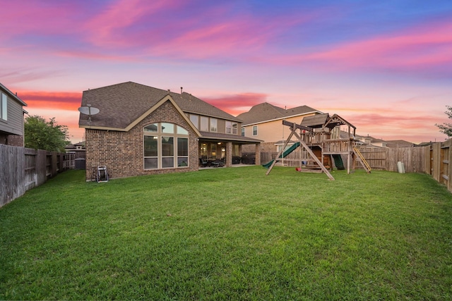 back of property with a fenced backyard, brick siding, roof with shingles, a playground, and a lawn