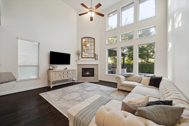 living room featuring a fireplace, a ceiling fan, baseboards, a towering ceiling, and dark wood-style floors