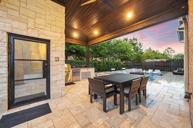 patio terrace at dusk featuring ceiling fan, a fenced backyard, an outdoor kitchen, outdoor dining space, and a grill