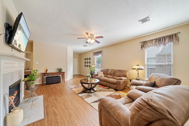 living area with a ceiling fan, light wood-type flooring, a tile fireplace, baseboards, and visible vents