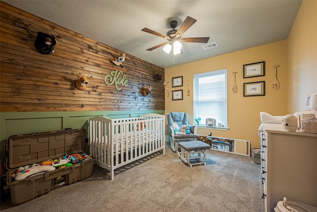bedroom with carpet floors, a crib, wood walls, visible vents, and a ceiling fan