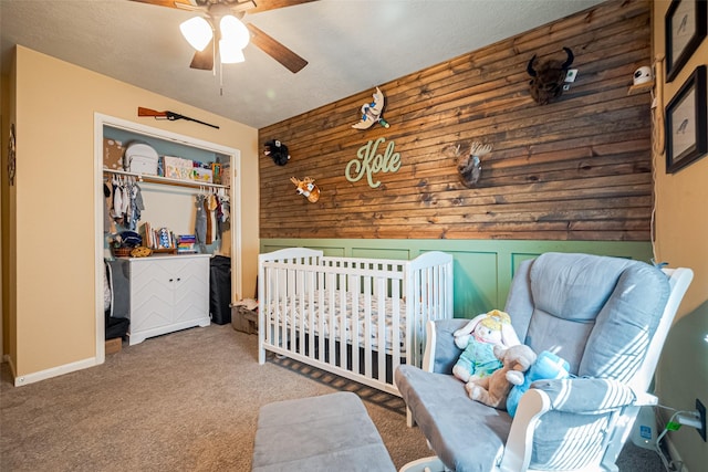 bedroom with wood walls, ceiling fan, a nursery area, carpet floors, and a closet