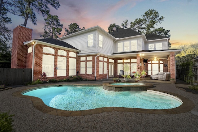 back of property at dusk featuring brick siding, a patio area, a pool with connected hot tub, and fence