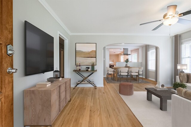 living area featuring light wood finished floors, arched walkways, baseboards, a ceiling fan, and ornamental molding
