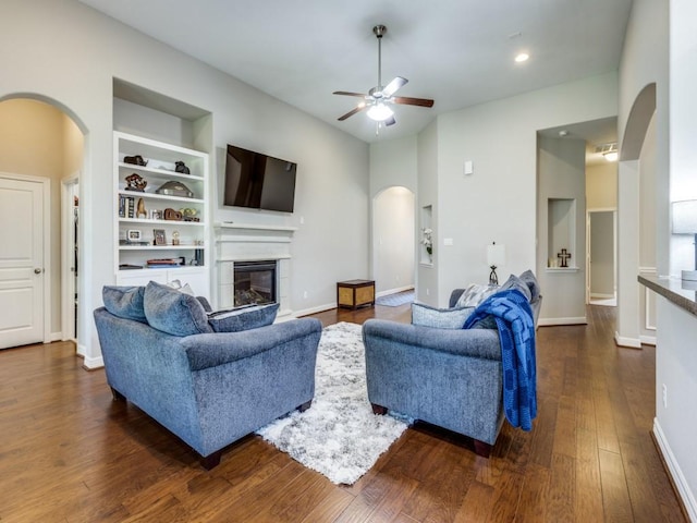living area with a glass covered fireplace, dark wood-style floors, a ceiling fan, arched walkways, and built in shelves